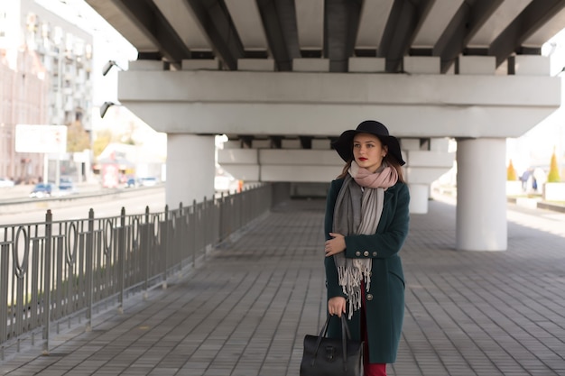 Urban portrait of young brunette woman wearing coat, black hat and purse. Space for text