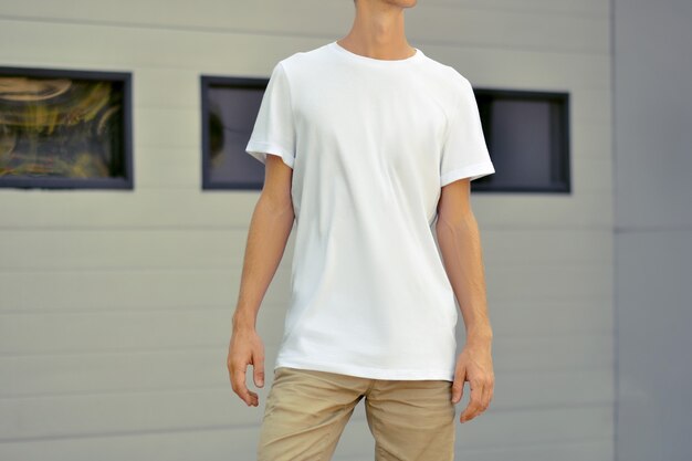 Urban template  of  clothes. Yong slim guy in a blank T-shirt and brown shorts stand near the white metal profiled wall with black windows. Mockup  can use for you design.