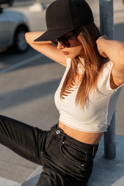 Urban young hipster girl in sunglasses in fashionable T-shirt in black American cap and jeans is resting in sun in city parking lot on summer day.