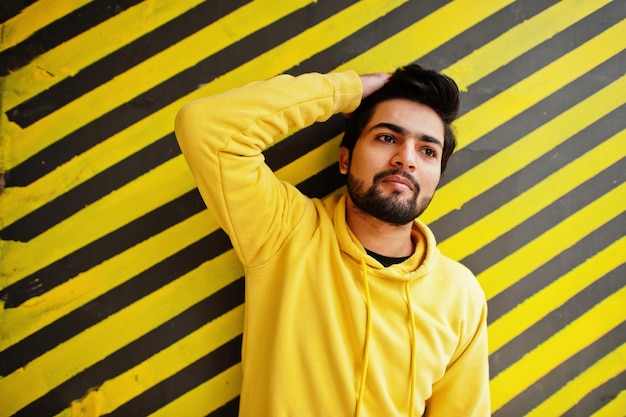 Urban young hipster indian man in a fashionable yellow sweatshirt Cool south asian guy wear hoodie against striped background