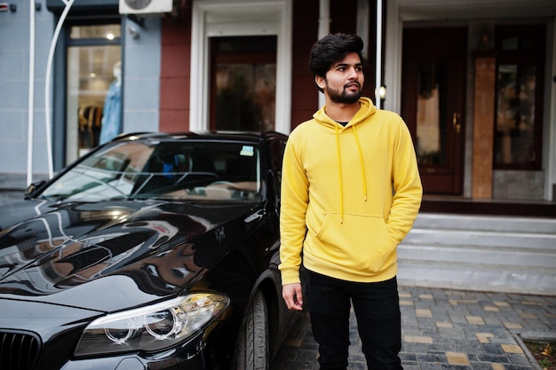 Urban young hipster indian man in a fashionable yellow sweatshirt Cool south asian guy wear hoodie stand on porch of the house with black business car