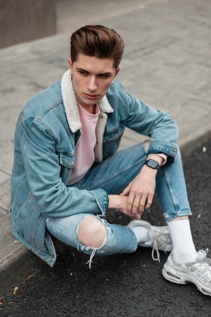 Urban young man model with hairstyle in fashionable blue denim jacket in stylish ripped jeans in white shoes rest on tile near road. Sweet guy in casual youth clothes on street in city. Summer style.