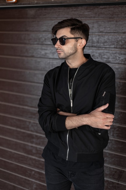 Urban young man in a stylish black jacket in trendy sunglasses in a fashionable hairstyle relaxes near a vintage brown wooden wall on the street. Attractive guy posing indoors. Street style.