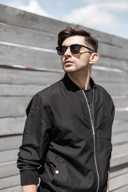 Urban young man in a stylish black jacket in trendy sunglasses in a fashionable hairstyle relaxes near a vintage wooden fence on a bright sunny day. Attractive guy enjoys the sunshine outdoors