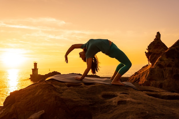 우르드바 다누라사나(Urdhva Dhanurasana) 바다의 등대 옆에 있는 일몰에 바위 위에서 명상과 요가 운동을 하는 여성 건강하고 자연주의적인 삶