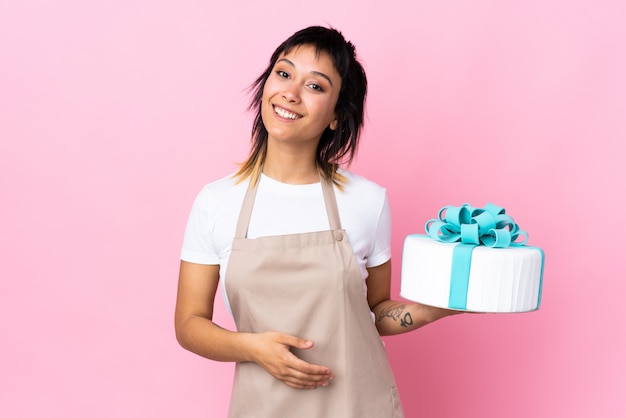 Uruguayaanse banketbakker die een grote cake over roze muur houdt die lege ruimte denkbeeldig op de palm houdt