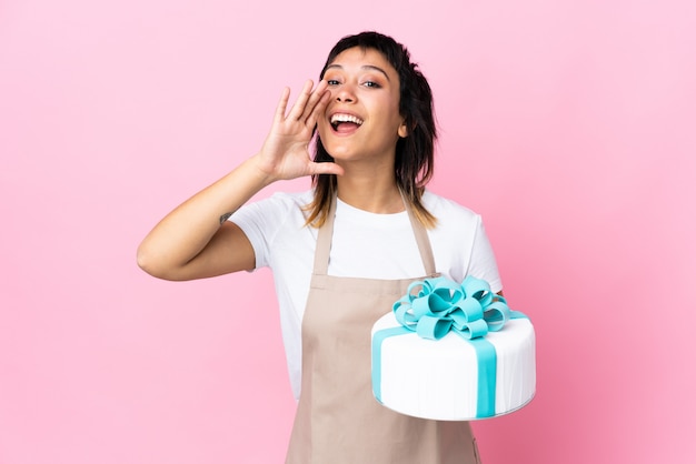 Uruguayan Pastry chef holding a big cake over isolated pink shouting with mouth wide open