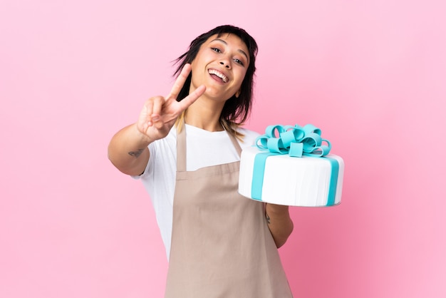 Uruguayan Pastry chef holding a big cake over isolated pink smiling and showing victory sign