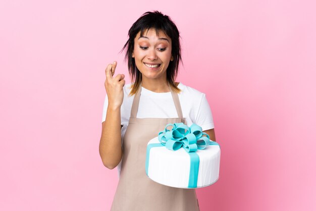 Cuoco unico di pasticceria uruguaiano che tiene una grande torta sopra il rosa isolato con l'incrocio delle dita