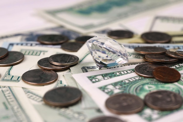 큰 다이아몬드가 있는 미국 화폐 지폐와 동전은 탁자 위에 있는 많은 양의 달러와 거대한 투명한 보석을 닫습니다.