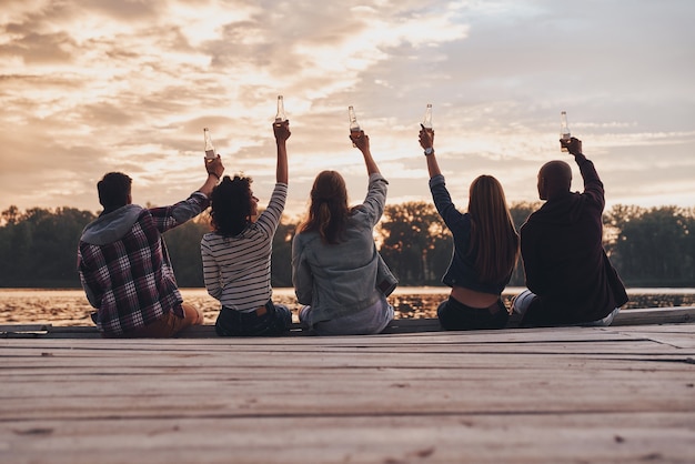 To us! Rear view of young people in casual wear toasting with a beer bottles 