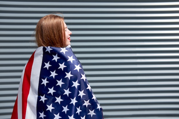 USA stars and stripes flag Young redhead woman with red painted lips standing with USA flag Grey metal panel background