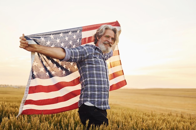 USA vlag in handen houden Patriottische senior stijlvolle man met grijs haar en baard op het landbouwgebied