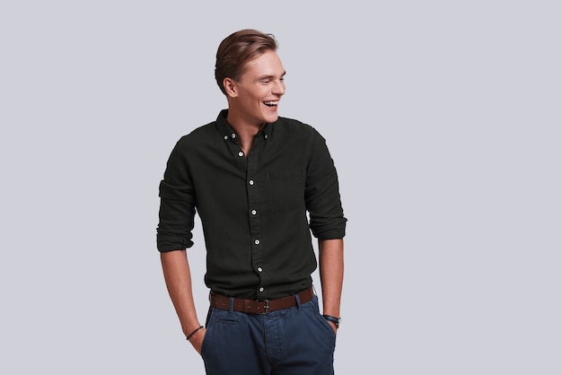 Used to look perfect. Good looking young man keeping hands in pockets and laughing while standing against grey background