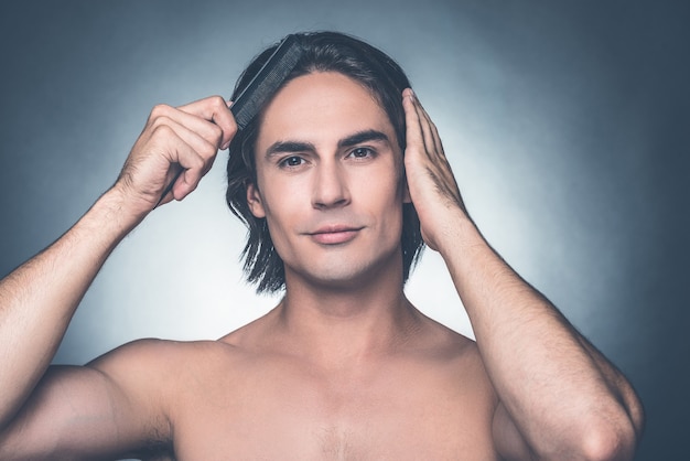 Used to look perfect. Portrait of young shirtless man combing his hair with hairbrush and looking at camera while standing against grey background