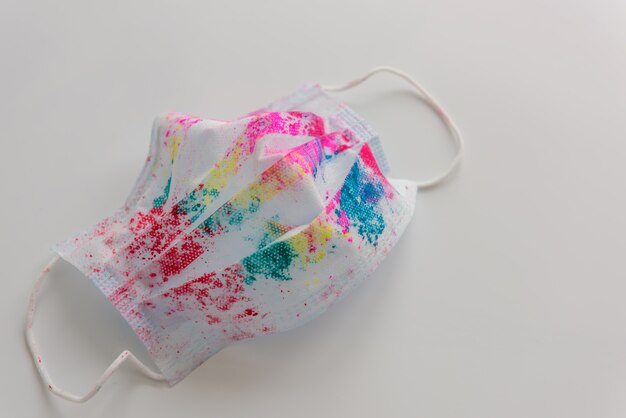 Used medical face mask with bright multicolored spots from holi paints, on white background, close-up. Concept of celebrating the Indian Happy Holi Festival during coronavirus pandemic. Copy space.