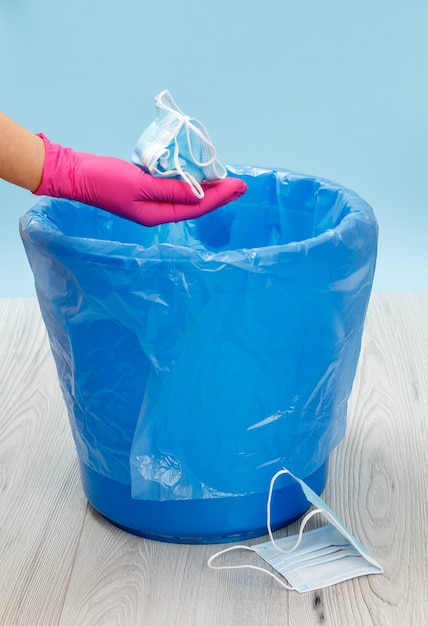 Used medical protective masks. The trash bin for utilization of individual protection means during of pandemic. Hand in a nitrile glove and a blue trash bag. Pollution of environment.