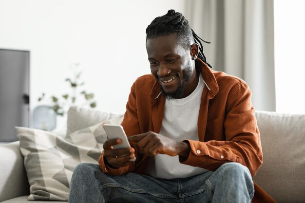 Useful application Happy africam american man using smatphone while resting on sofa at home copy space