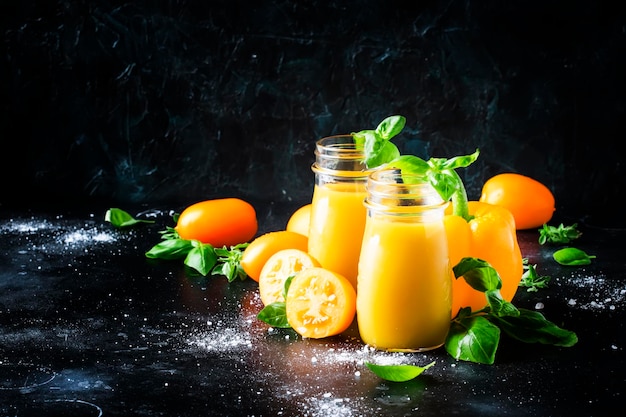 Useful and healthy smoothie or juice from yellow tomatoes and bell peppers with green basil in glass bottles black background selective focus