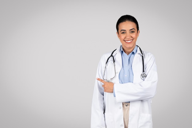 Useful information portrait of cheerful latin female doctor pointing at copy space grey background