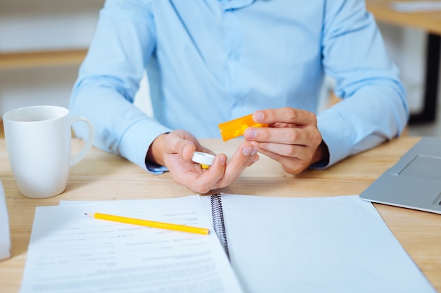 Useful pills. Serious office worker sitting in front of camera and wearing blue shirt while taking tablets