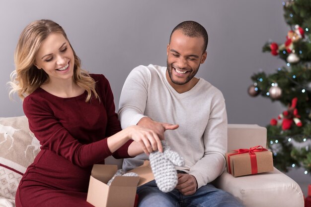 Useful present. Joyful handsome positive man laughing and trying on a mitten while being helped by his wife