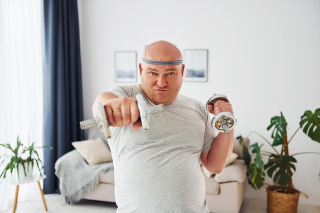 Photo uses dumbbells funny overweight man in casual clothes is indoors at home