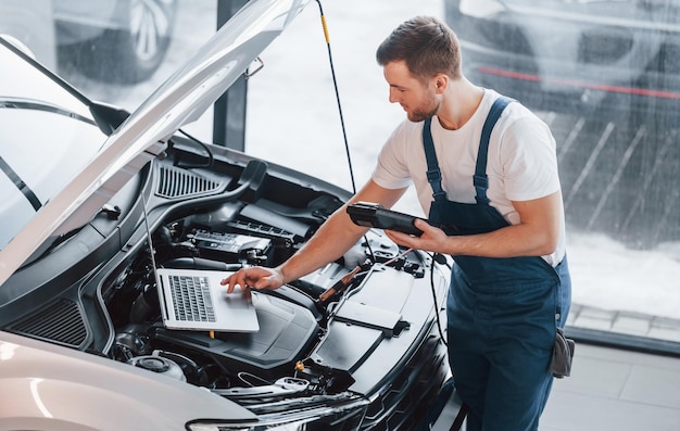 Uses laptop Young man in white shirt and blue uniform repairs automobile