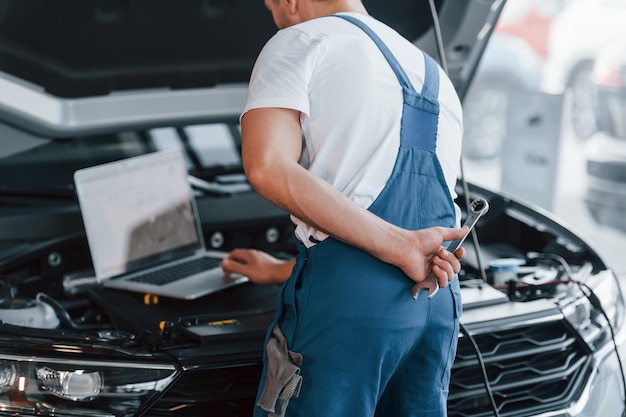 Использует ноутбук Молодой человек в белой рубашке и синей форме ремонтирует автомобиль