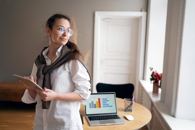 Uses a tablet a woman with glasses looks at the camera in a white shirt sitting in the office