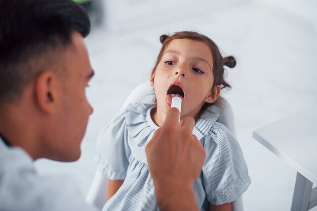 Uses tonsil to check throat. Young pediatrician works with little female visitor in the clinic.