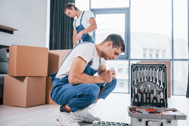 Uses tools from case Two young movers in blue uniform working indoors in the room