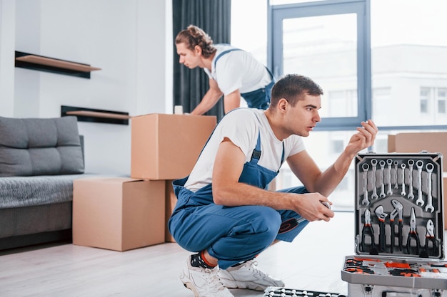 Uses tools from case Two young movers in blue uniform working indoors in the room