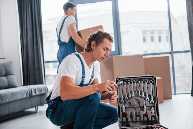 Uses tools from case two young movers in blue uniform working\
indoors in the room