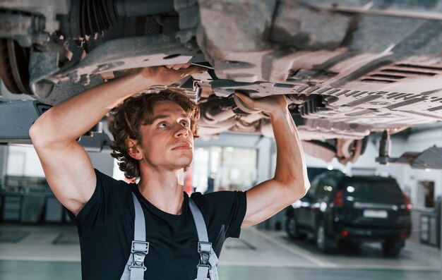 Uses wrench to fix car adult man in grey colored uniform works\
in the automobile salon