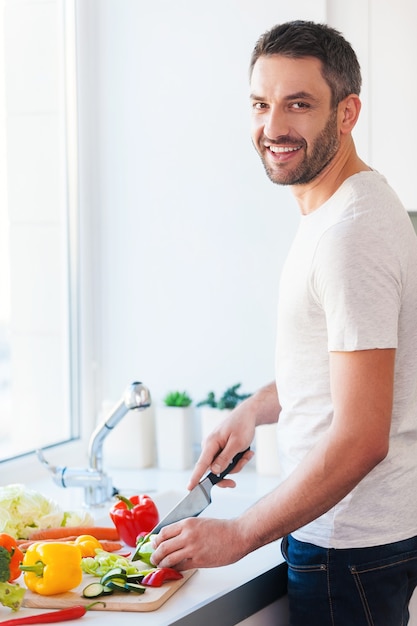 Usando le migliori verdure per la mia insalata. bel giovane che taglia le verdure e sorride mentre sta in cucina