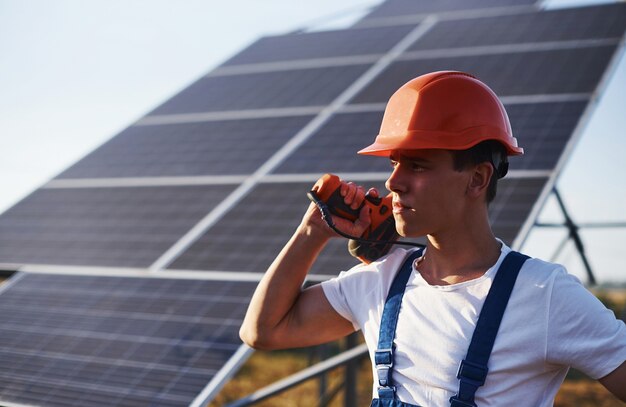 Using cordless screwdriver. Male worker in blue uniform outdoors with solar batteries at sunny day.