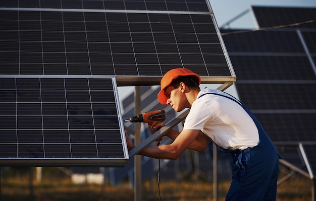 С помощью аккумуляторной отвертки. Работник-мужчина в синей форме на открытом воздухе с солнечными батареями в солнечный день.