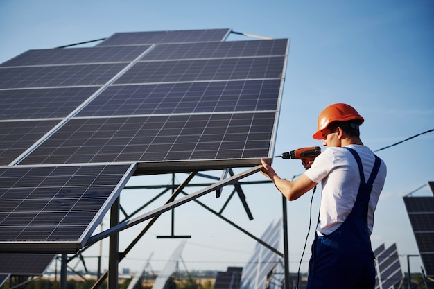 Using cordless screwdriver. Male worker in blue uniform outdoors with solar batteries at sunny day.