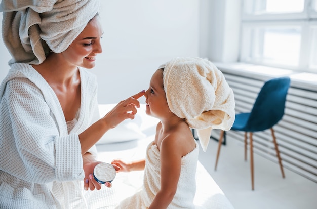 Foto usando la crema per pulire la pelle. la giovane madre con sua figlia ha una giornata di bellezza al chiuso nella stanza bianca.