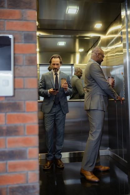 Using elevator. Businessmen wearing suits holding coffee using elevator in business center