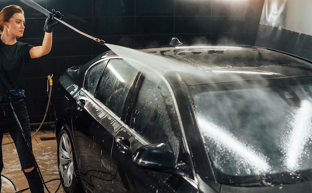 Utilizzo di apparecchiature con acqua ad alta pressione la moderna automobile nera viene pulita da una donna all'interno della stazione di lavaggio auto