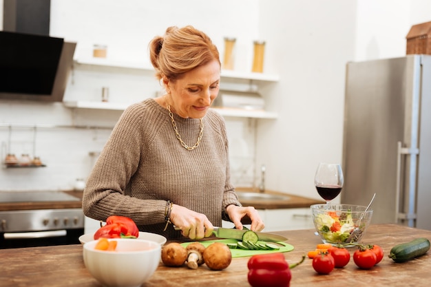 新鮮な野菜を使用。金属ナイフを運んでいる間、木製のテーブルで夕食の準備をしている集中した成熟した女性