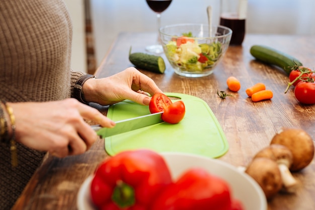 Using fresh vegetables. Pleasant woman carrying long metal knife and chopping tomato on green board