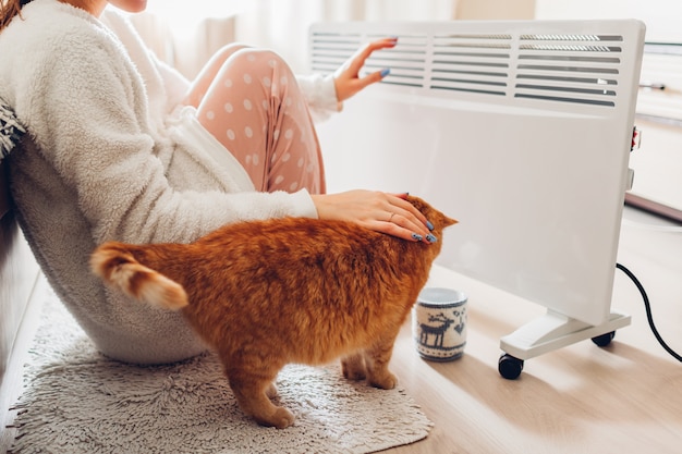 Using heater at home in winter. Woman warming her hands with cat. Heating season.