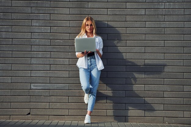 Using laptop Beautiful blonde in casual clothes is outdoors at sunny daytime