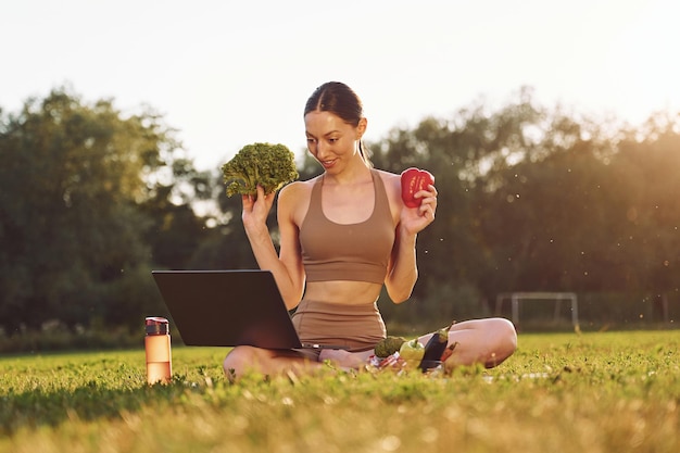 Foto utilizzo di laptop tenere verdure giovane donna in abiti da yoga è all'aperto sul campo