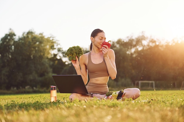 Использование ноутбука Холдинг овощей Молодая женщина в одежде для йоги находится на открытом воздухе на поле