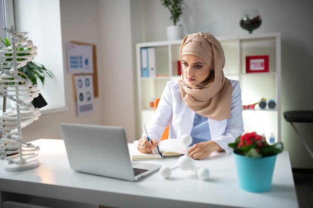 Using laptop. Medical scientist wearing headscarf working using laptop and making some notes