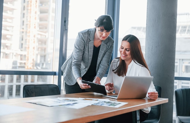 Using laptop Two women in formal clothes is indoors in the modern office works together